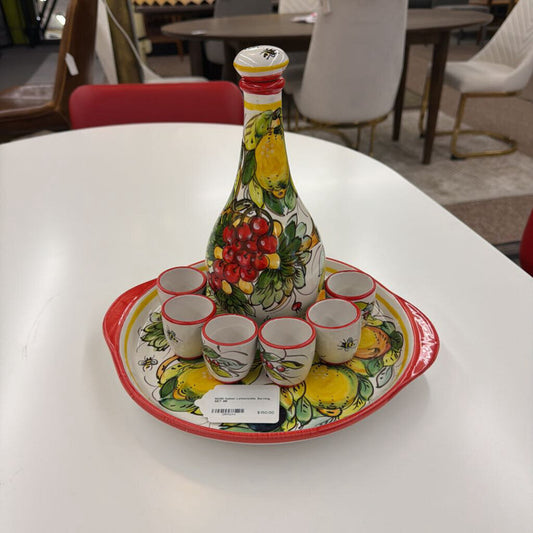 Decorative ceramic set with a bottle and six cups on a tray, featuring fruit designs, on a white table.