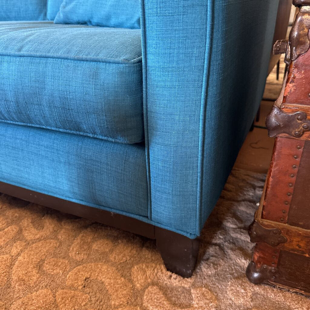 Close-up of a blue textured sofa corner with a brown leather suitcase on a patterned rug.