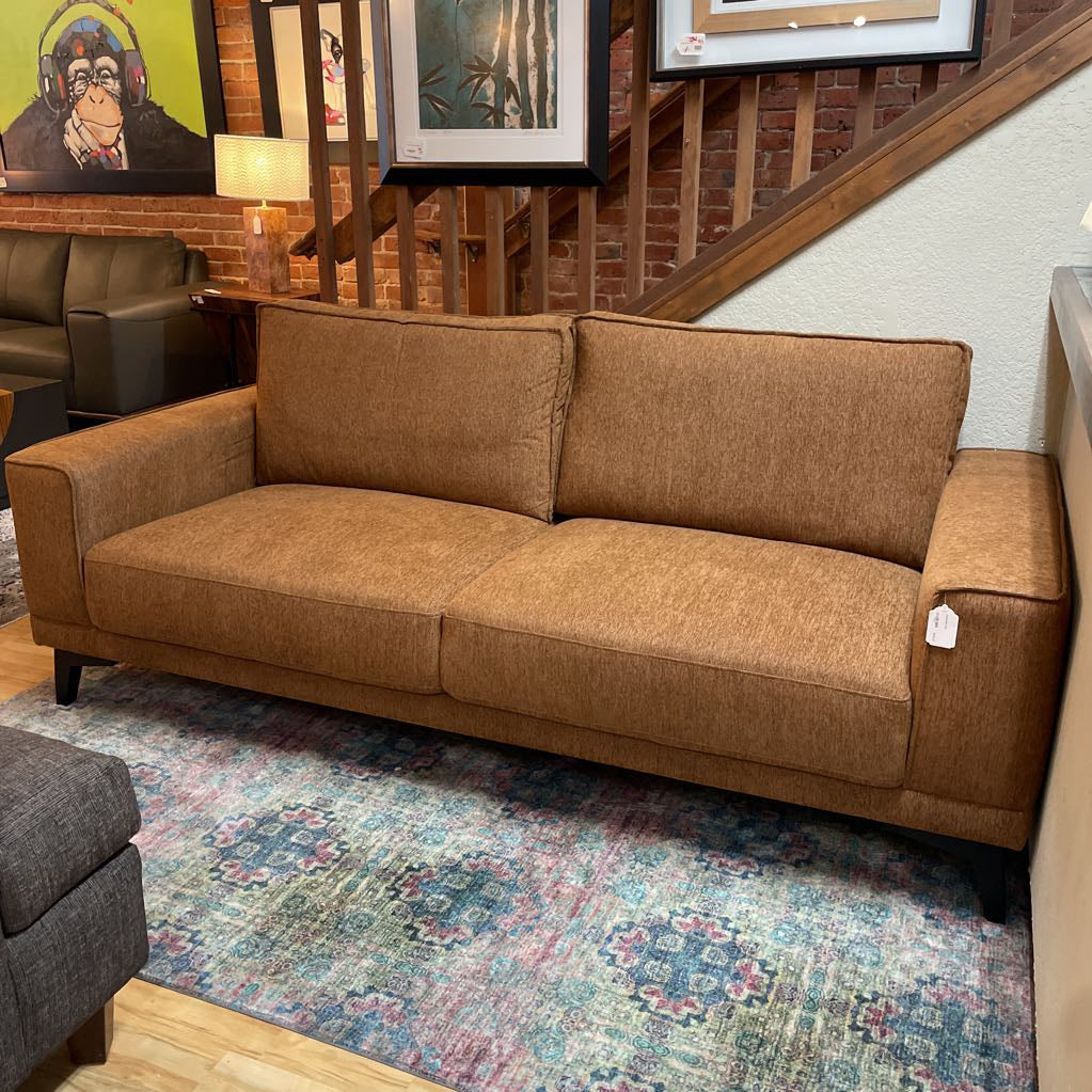 Brown sofa in a room with a colorful rug and framed pictures on the wall.
