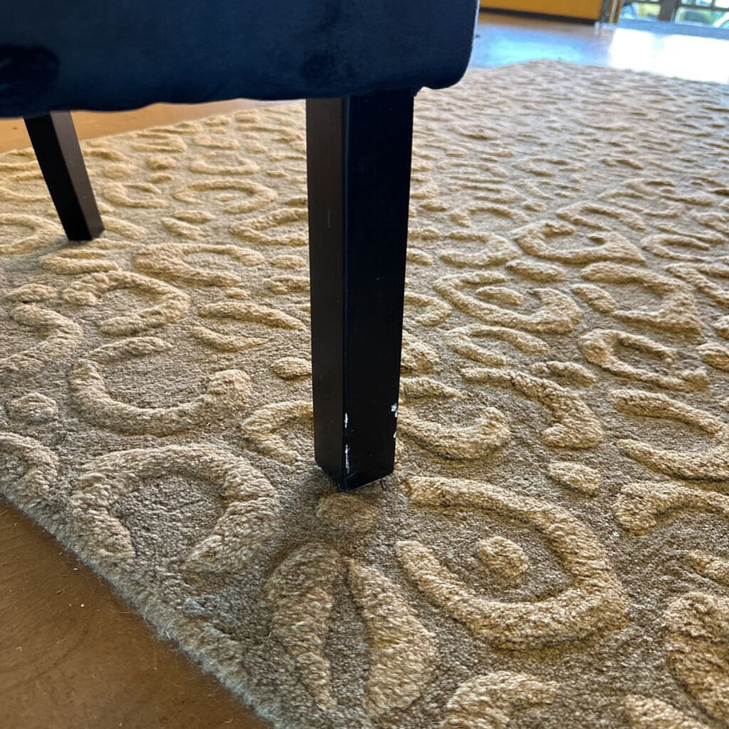 Close-up of a textured beige carpet with a dark blue chair leg in the foreground.
