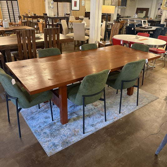 Wooden dining table with green chairs on a blue rug in a showroom setting.