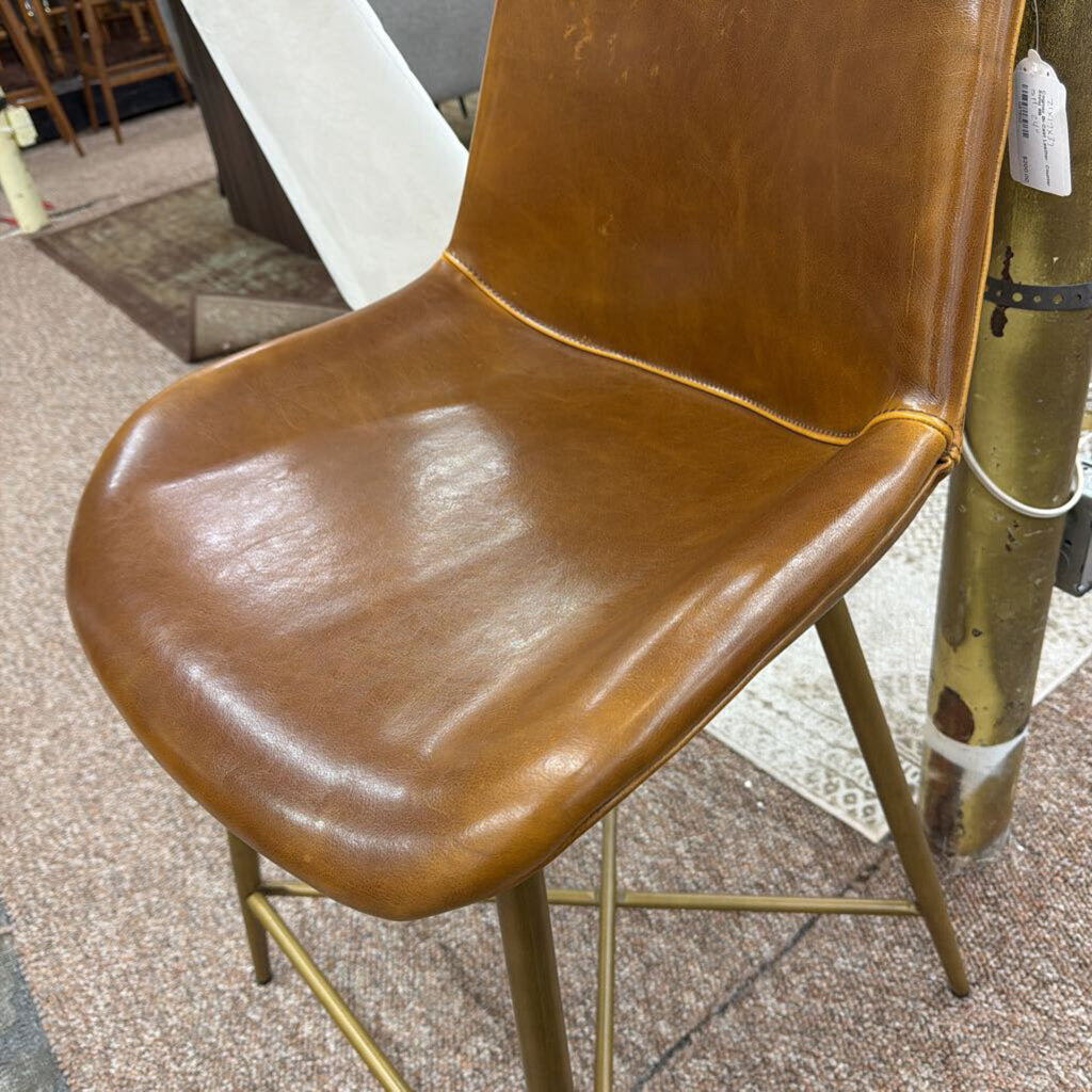 Brown leather chair with gold metal legs on a textured floor.