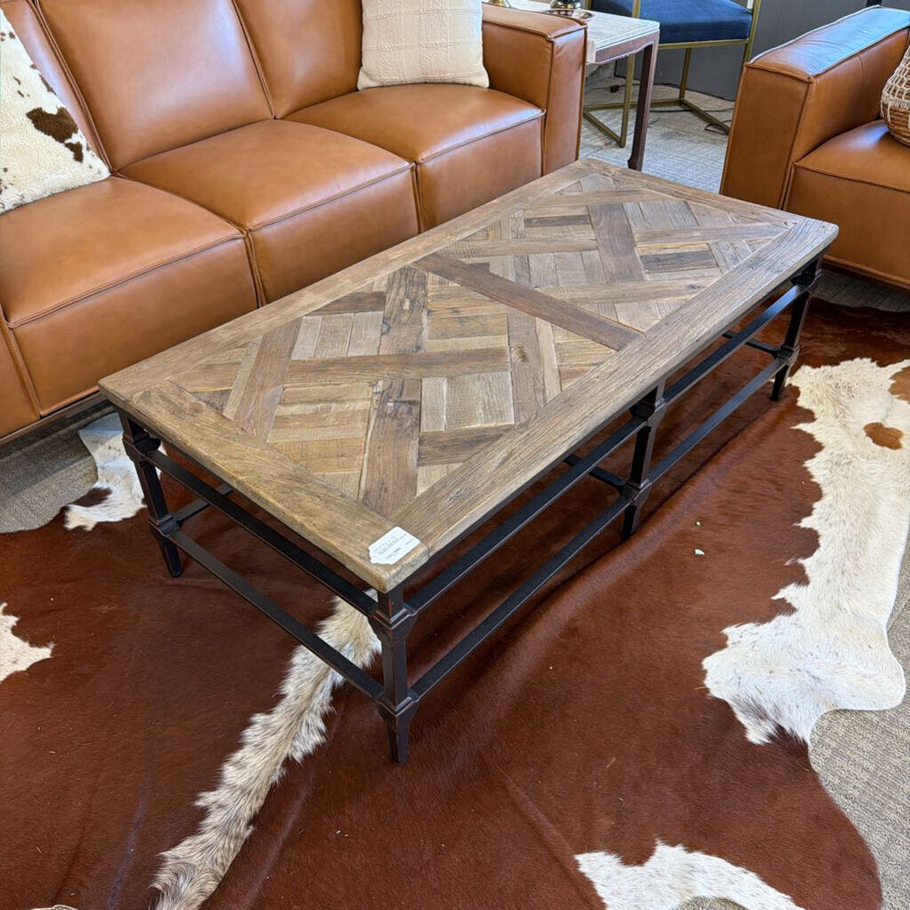Wooden coffee table with a herringbone pattern in a living room setting with brown leather sofa and cowhide rug.
