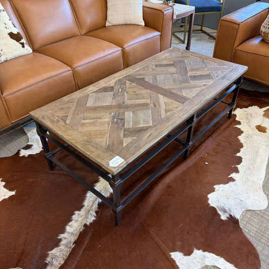 Wooden coffee table with a herringbone pattern in a living room setting with brown leather sofa and cowhide rug.