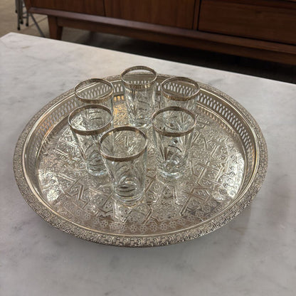 Set of six clear glasses on an ornate silver tray on a marble surface.
