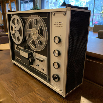 Vintage reel-to-reel tape recorder on a wooden floor with a blurred indoor background