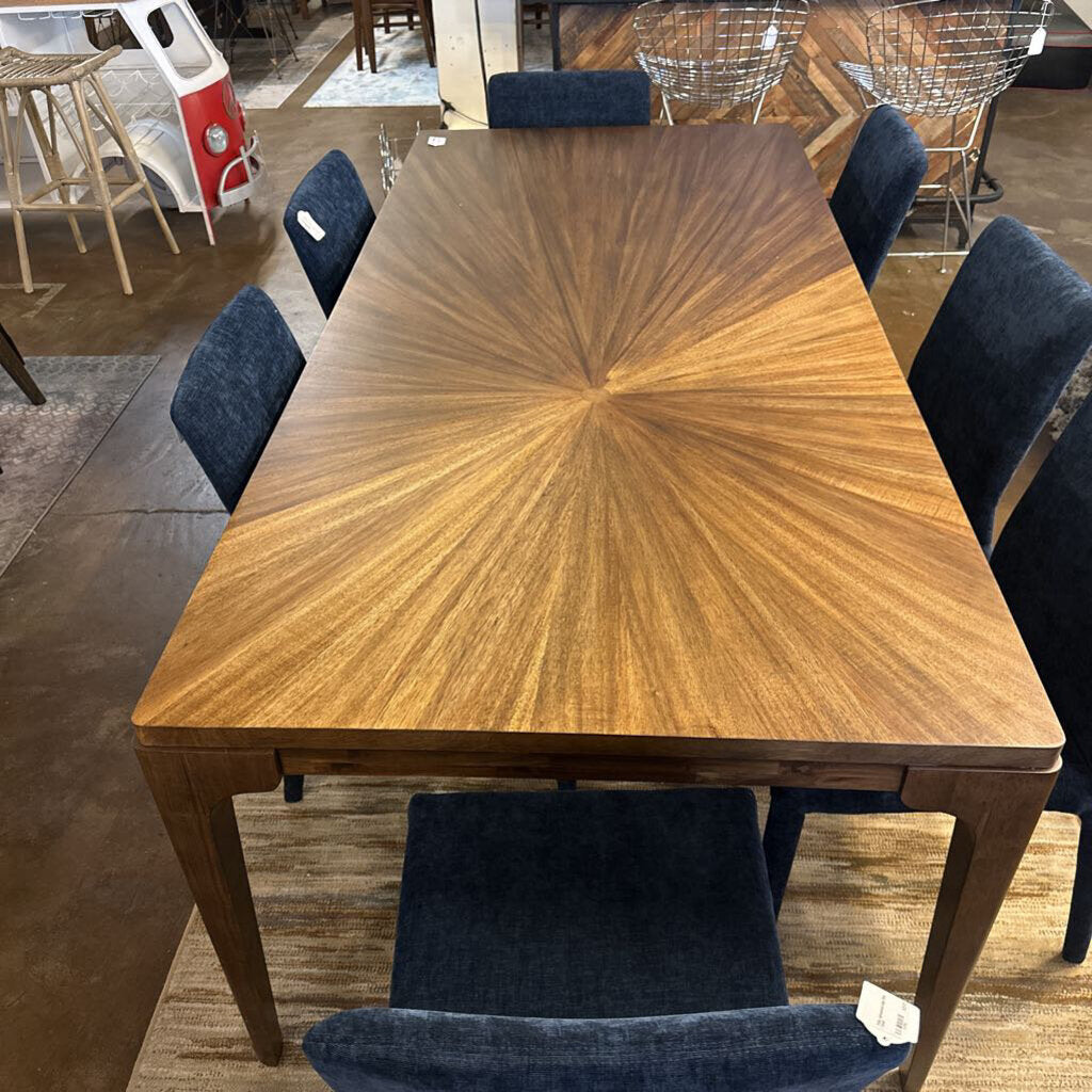 Wooden dining table with starburst pattern surrounded by dark blue chairs in a showroom setting.