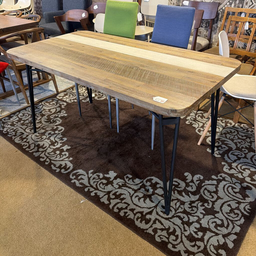 Wooden dining table with metal legs on a patterned rug in a room with chairs and tables.