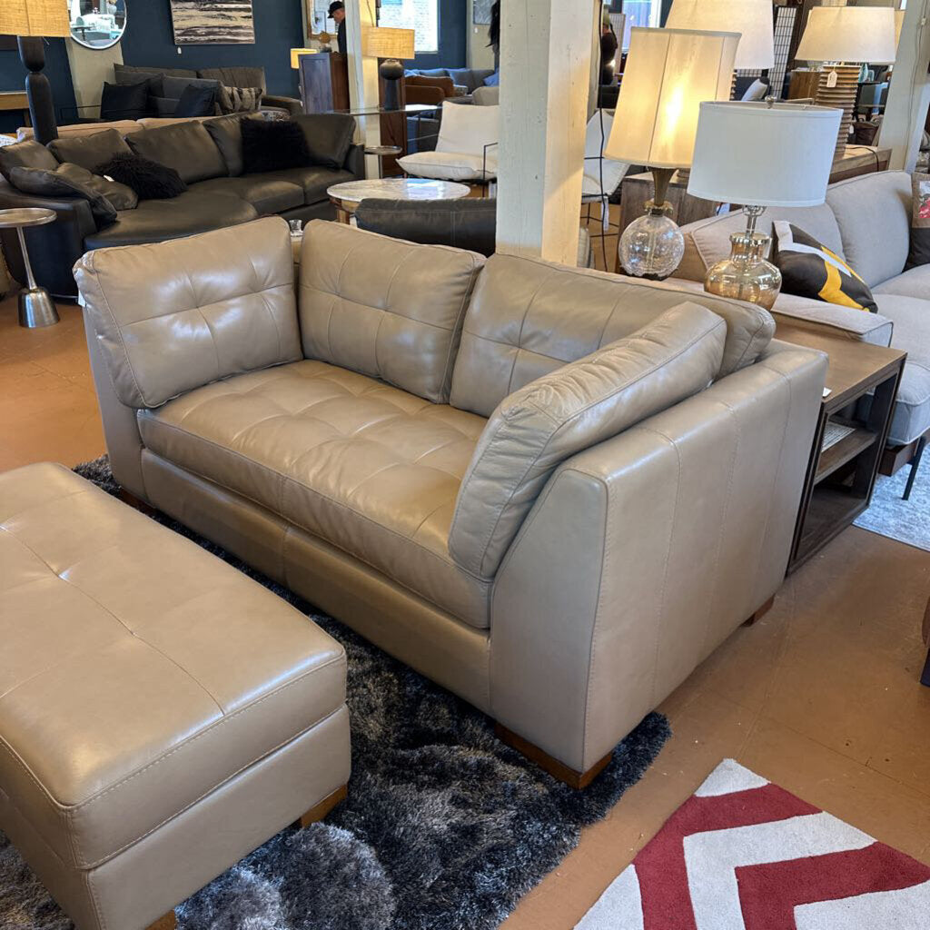 Beige leather sofa and ottoman in a showroom setting with other furniture.