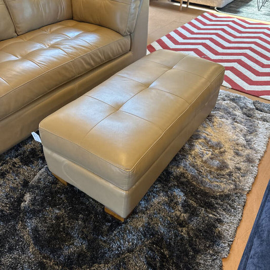 Beige ottoman on a dark textured rug with a patterned red and white rug in the background.