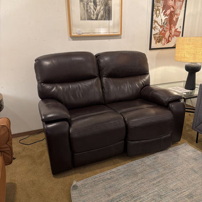 Brown leather recliner sofa in a living room setting with a lamp and framed pictures on the wall.