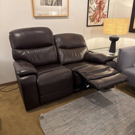 Brown leather recliner sofa in a living room setting with a lamp and framed pictures on the wall.