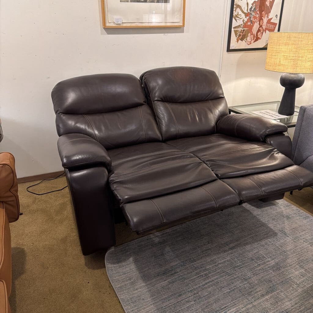 Brown leather recliner sofa in a room with a lamp and framed pictures on the wall.