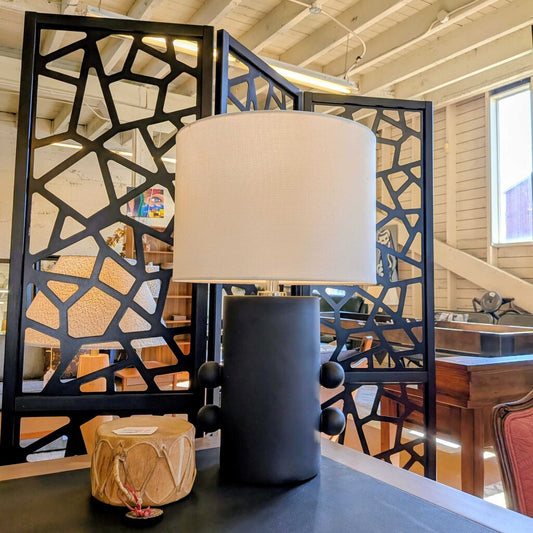 Decorative lamp with a black base and white shade in a room with geometric screen and wooden furniture.
