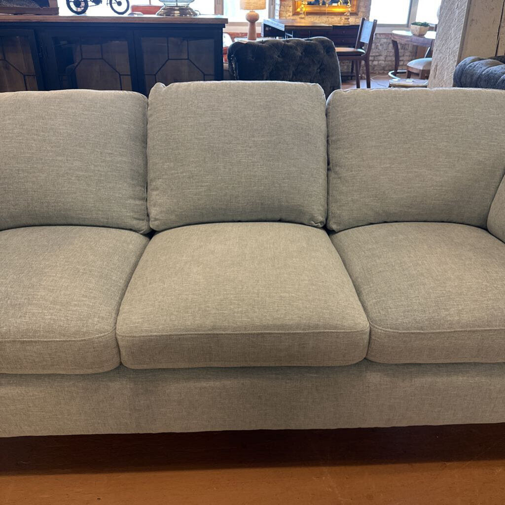 Beige sofa in a living room setting with furniture and decor in the background.