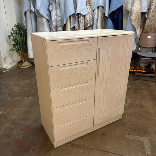 Wooden cabinet with drawers and a door in an indoor setting with clothes in the background.