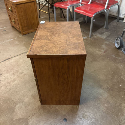 Wooden nightstand on a concrete floor with red chairs in the background
