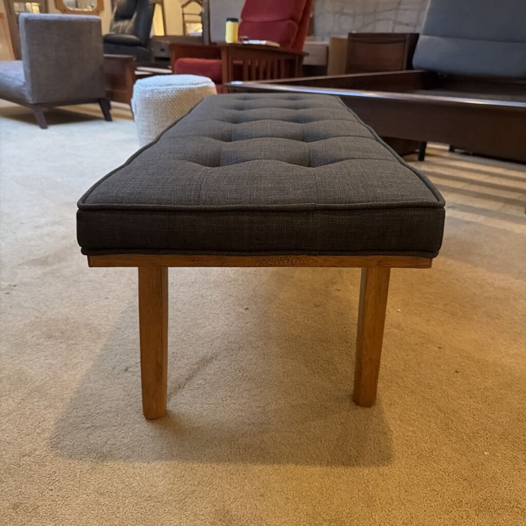 Gray cushioned ottoman on a carpeted floor with furniture in the background