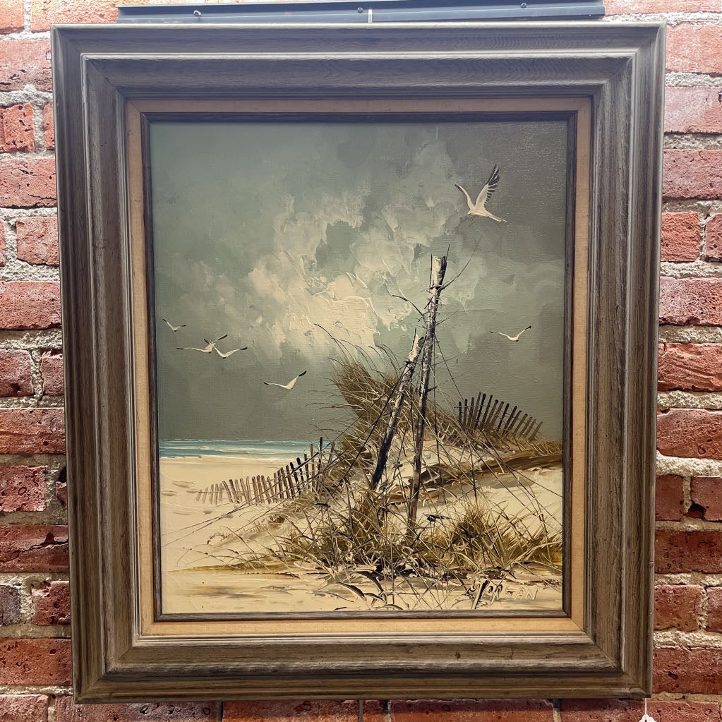 Framed painting of a beach scene with birds and a fence on a brick wall.