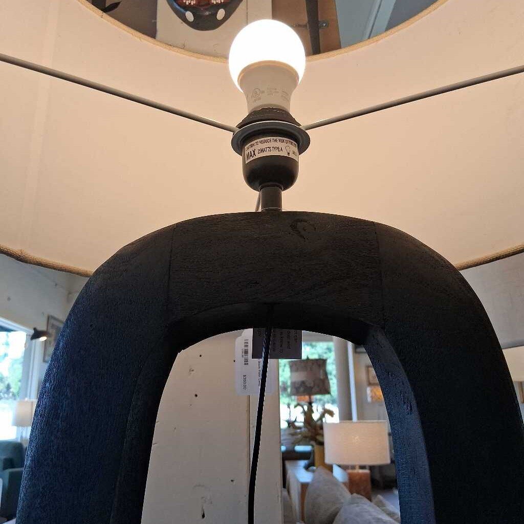 Close-up of a lamp with a black shade and visible bulb, with a blurred indoor setting in the background.