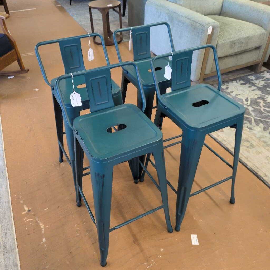 Set of teal metal bar stools with tags on a brown floor, furniture in the background