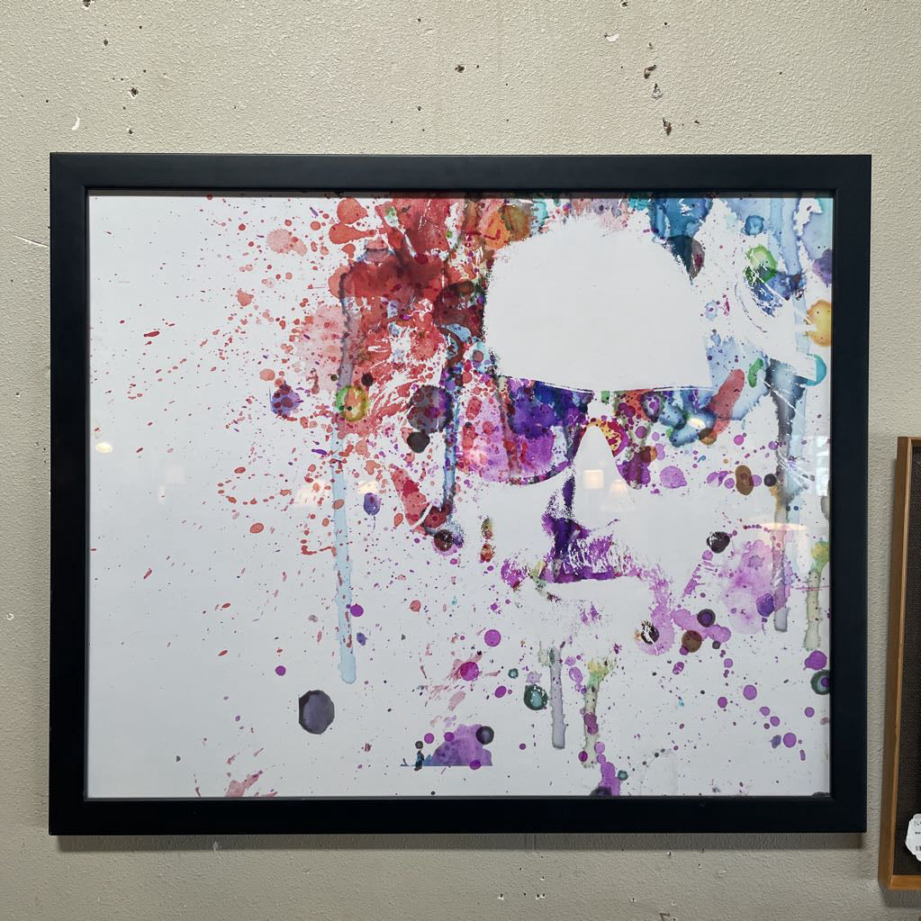 A framed watercolor-style portrait in a black frame, featuring a minimalist silhouette of a man with sunglasses created through vibrant purple, pink, and teal paint splatters and drips on a white background