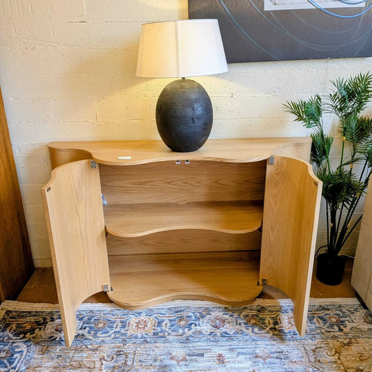 Wooden sideboard with open doors, lamp, and plant on a patterned rug.