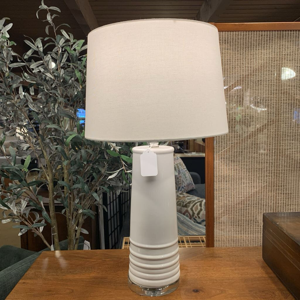 Beige table lamp with a white shade on a wooden surface, with a plant and blurred background.