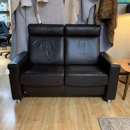 Black leather sofa in a showroom setting with a wooden floor and other furniture in the background.