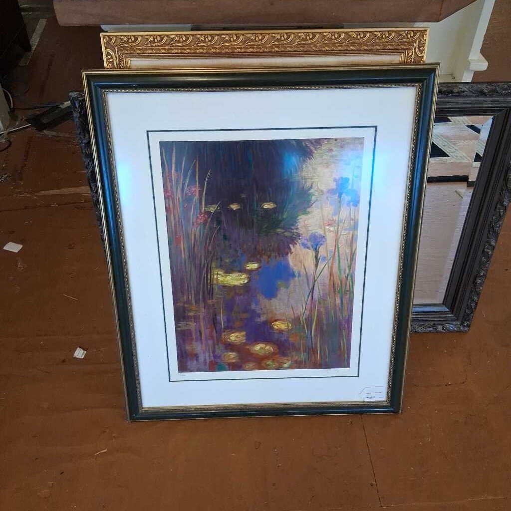 Framed artwork of a nature scene with flowers and water on a wooden surface.