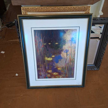 Framed artwork of a nature scene with flowers and water on a wooden surface.