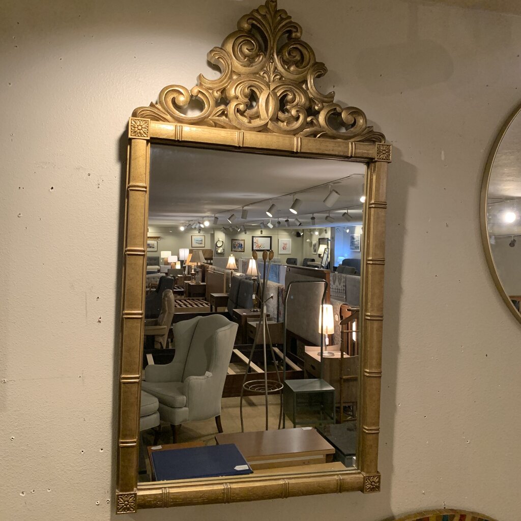 Decorative gold-framed mirror on a wall with a blurred indoor setting in the reflection.