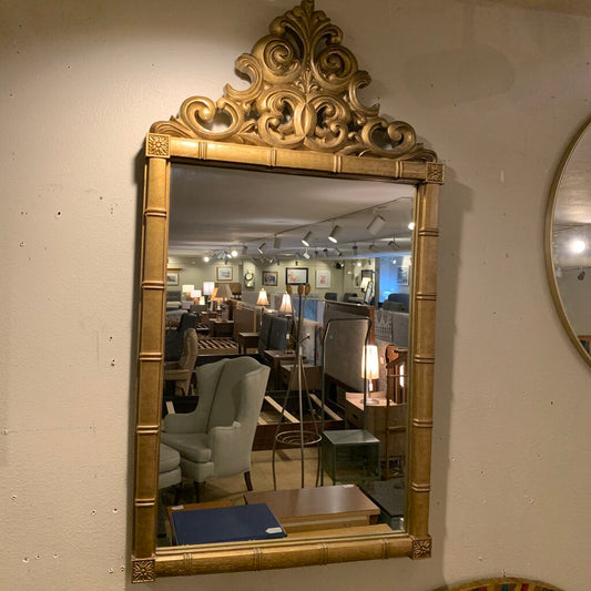 Decorative gold-framed mirror on a wall with a blurred indoor setting in the reflection.