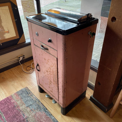 Vintage pink metal cabinet with black top on a wooden floor.