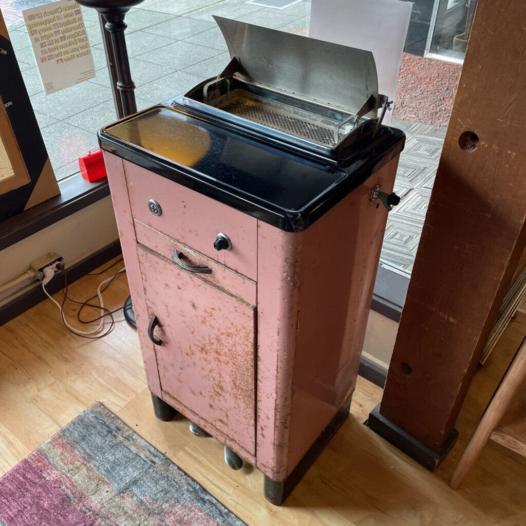 Pink vintage cabinet with a grill on top in a room with wooden floor and window.