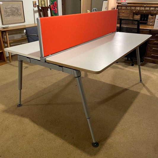 The desk comprises two off-white laminate work surfaces with a thin, natural wood-effect edge. Dividing the two stations is a vibrant orange fabric privacy screen that serves as a visual barrier and an acoustic damper. 