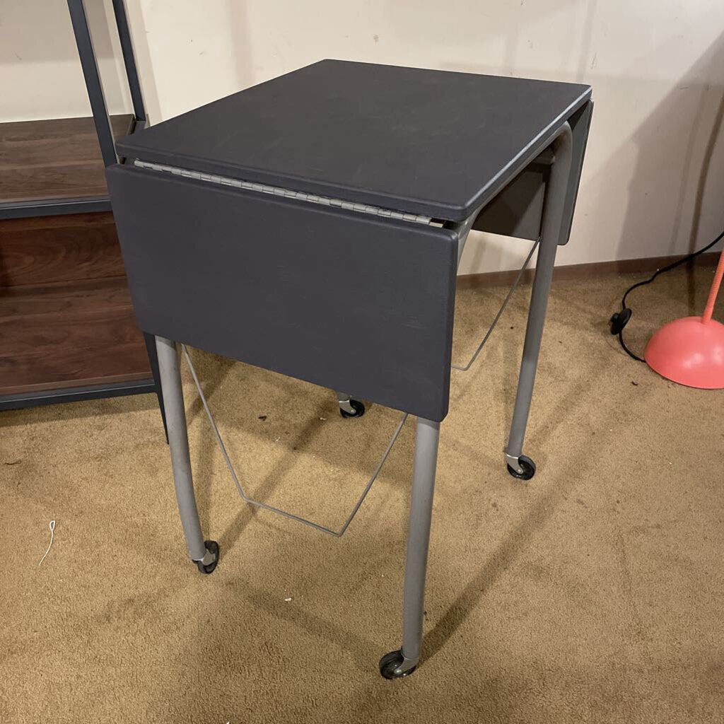 Gray metal folding table on wheels in a room with a carpeted floor.