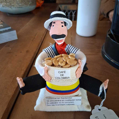 Figurine holding a bag of coffee labeled 'Cafe de Colombia' on a wooden surface.