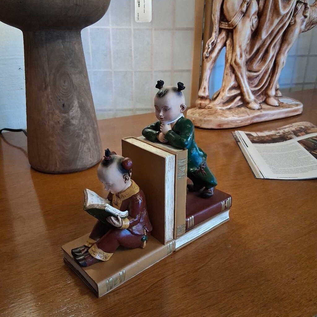 Two figurines on books on a wooden surface with a statue in the background.