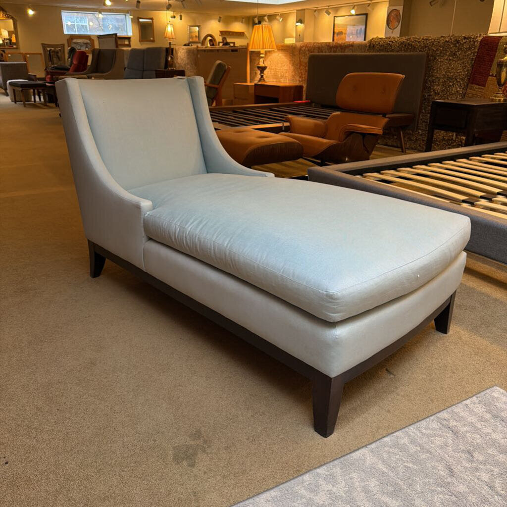 White upholstered chaise lounge in a showroom setting with furniture and decor in the background.