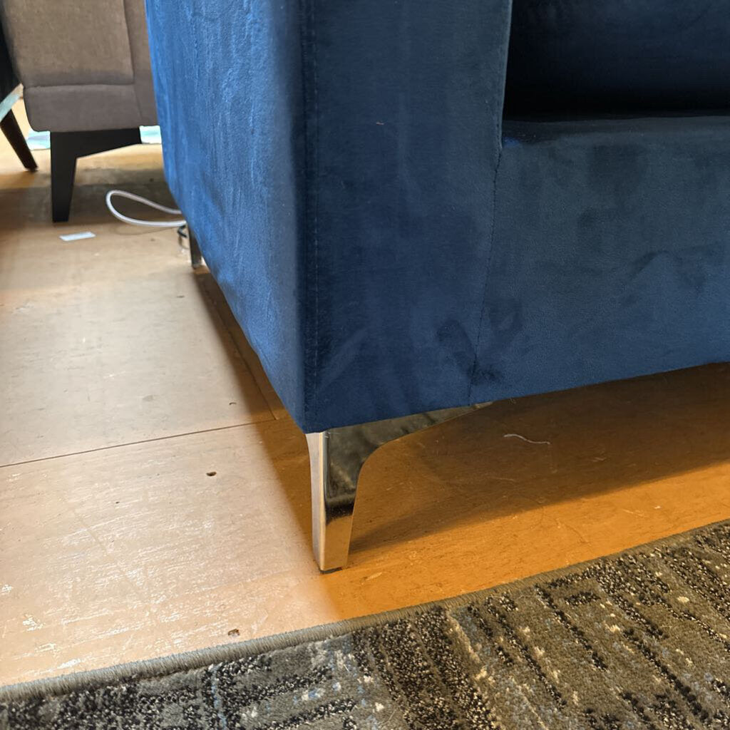 Blue velvet sofa with chrome legs on a wooden floor.
