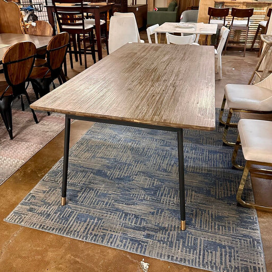 Wooden dining table with metal legs on a blue patterned rug in a showroom setting.