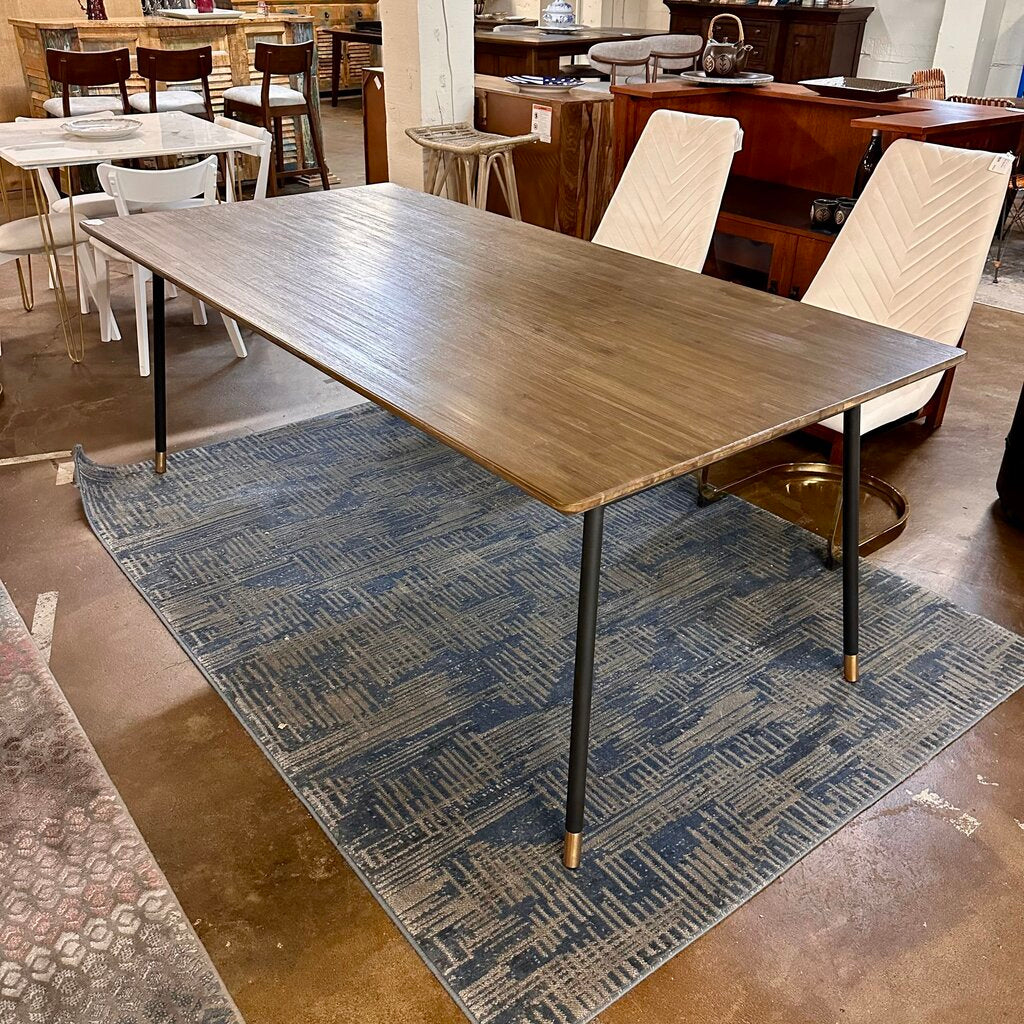 Wooden dining table with black legs on a blue patterned rug in a room with chairs and tables.