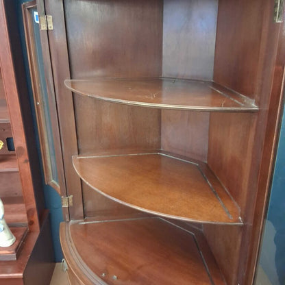Wooden corner cabinet with glass shelves in a room setting.