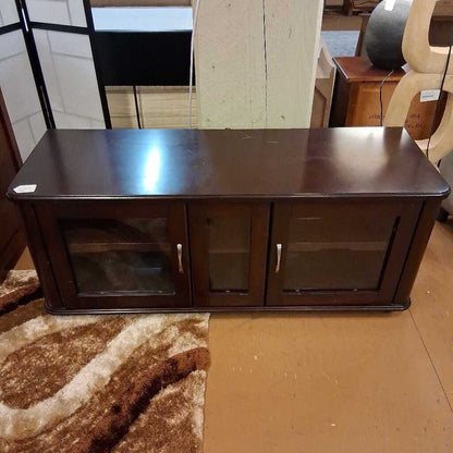 Dark wooden TV stand with glass panel and metal handles on a brown floor.