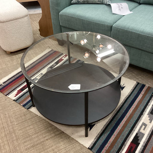 Round glass coffee table in a living room setting with a teal sofa and patterned rug.