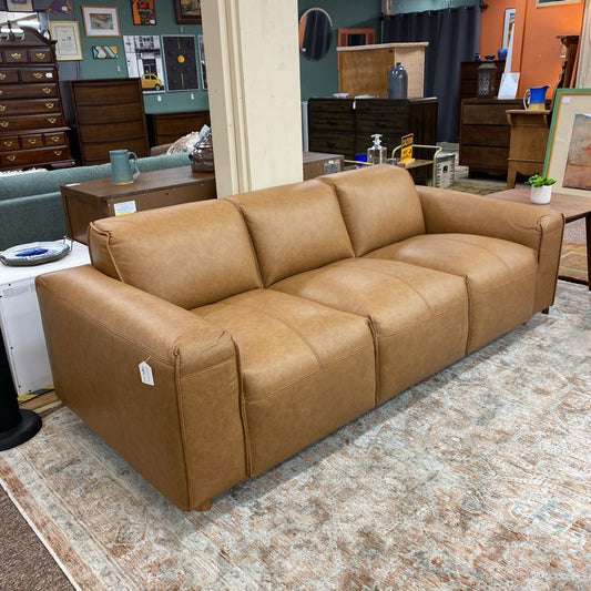 Brown leather sofa in a furniture store setting with various items in the background.