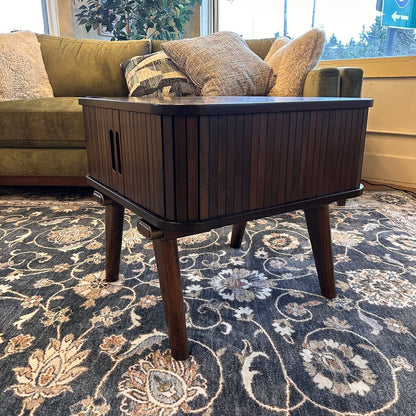 Wooden side table in a living room with a green sofa and patterned rug.