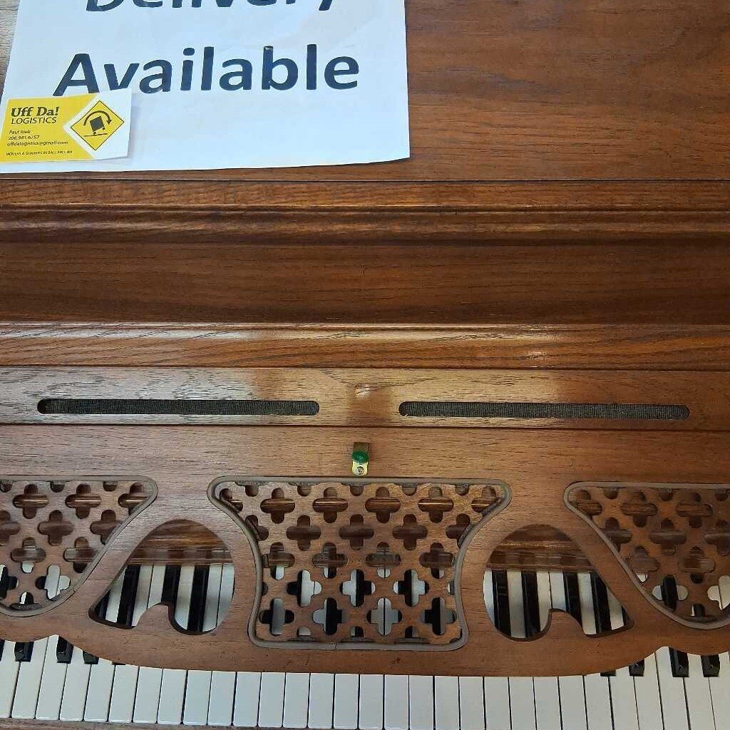 Wooden keyboard instrument with decorative carvings and a 'Delivery Available' sign above it.