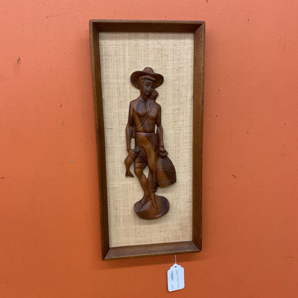 Wooden sculpture of a person with a hat and bag in a brown frame on an orange wall.